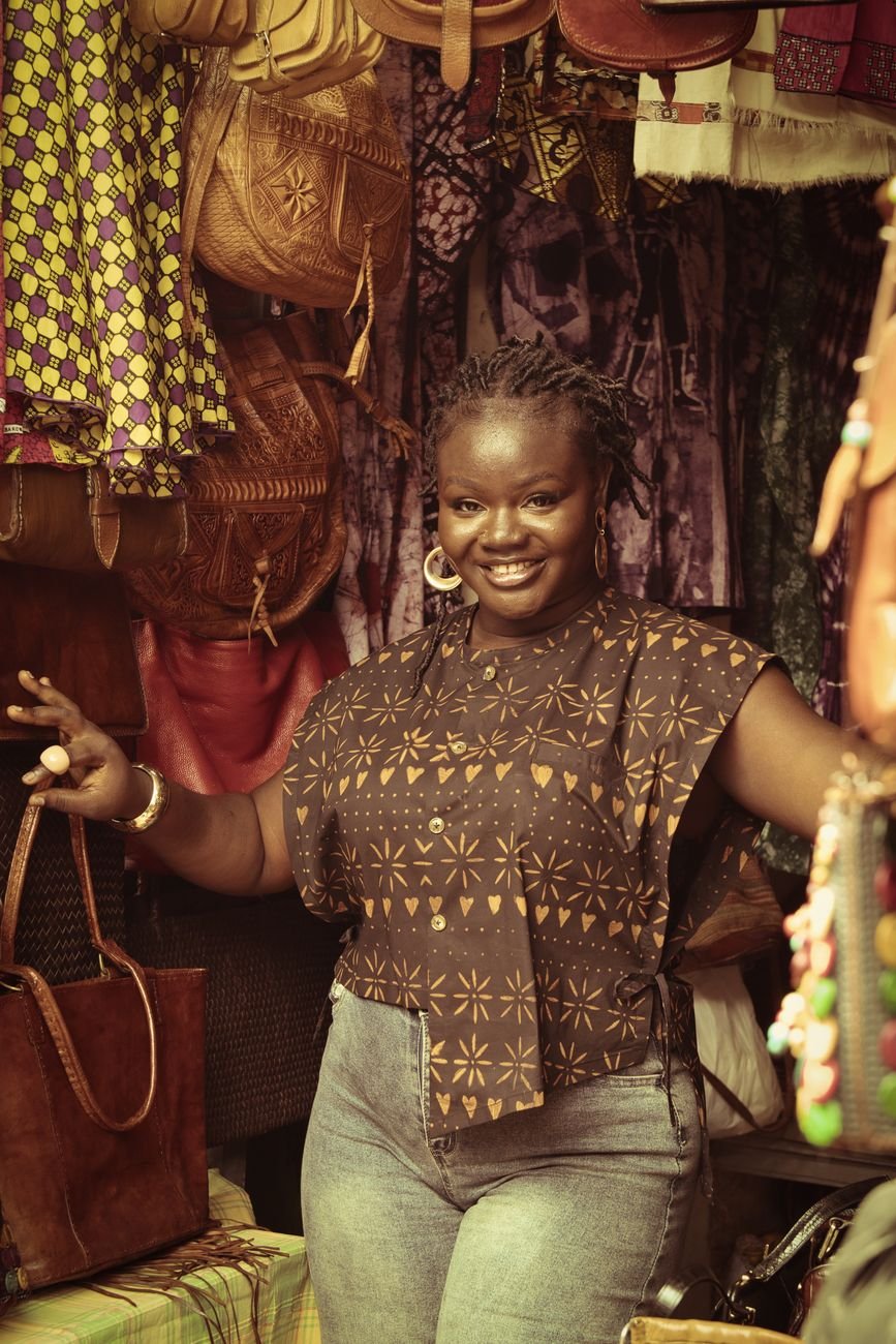 Smiling woman stands in colorful African market stall filled with handmade products.
